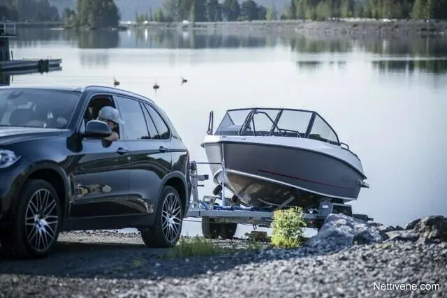 A black SUV parked near a lake, towing a boat on a trailer, with trees and ducks visible in the calm water.